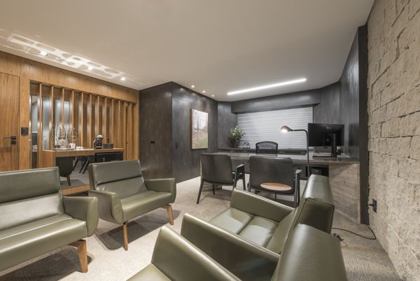 Poltronas de couro verde, painel ripado em madeira, aparador com frigobar e mesa de trabalho ao fundo